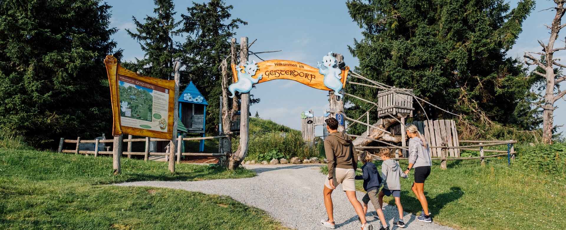 Familie mit zwei Kindern betritt bei Sonnenschein den fantasievollen Eingang zum Geisterberg mit Holzschild und Geisterfiguren