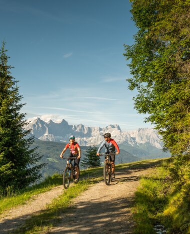 Zwei Mountainbiker fahren auf einem Waldweg mit Blick auf das Dachsteinmassiv, umgeben von grünen Bäumen und Wiesen. | © Altenmarkt-Zauchensee Tourismus/Lorenz Masser