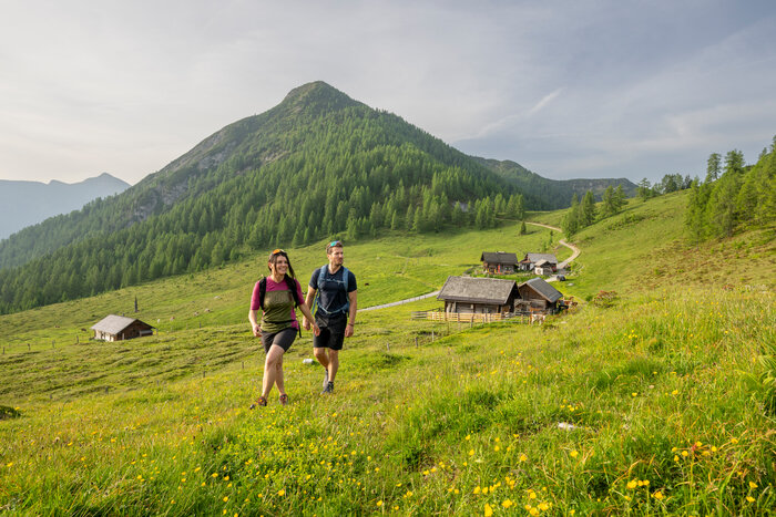 Zwei Wanderer gehen über eine Almwiese bei Altenmarkt, vor Holzhütten und einem bewaldeten Berg. | © Lorenz Masser