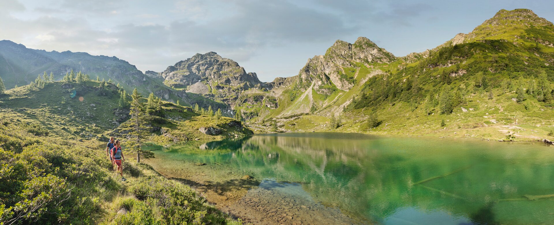 Zwei Wanderer gehen entlang eines türkisgrünen Bergsees, umgeben von Almwiesen, Bäumen und felsigen Bergen | © Peter Burgstaller