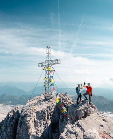 Zwei Kletterer mit Helmen stehen neben einem Gipfelkreuz auf felsigem Berggipfel über weiter Alpenlandschaft | © Mathäus Gartner