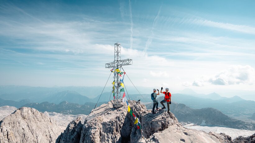 Zwei Kletterer mit Helmen stehen neben einem Gipfelkreuz auf felsigem Berggipfel über weiter Alpenlandschaft | © Mathäus Gartner