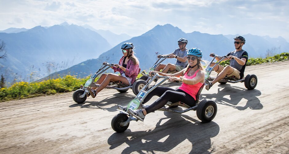 Vier Personen mit Helmen fahren auf Mountain Go-Karts eine breite Schotterstraße entlang, Berge im Hintergrund | © Planai, Harald Steiner