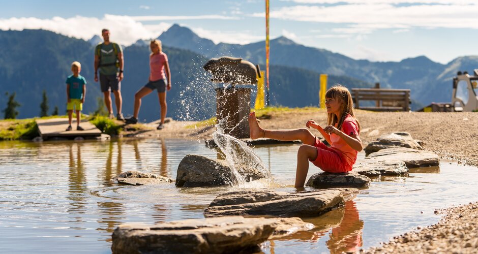 Ein Mädchen sitzt auf Steinen im flachen Wasser und spritzt, während eine Familie vor Bergkulisse steht | © Planai, Christine Höflehner