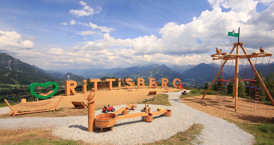 Panorama-Spielplatz mit Holzbuchstaben, Schaukeln und Wasserspiel vor Bergkulisse am Rittisberg | © Photo Austria, Hans-Peter Steiner