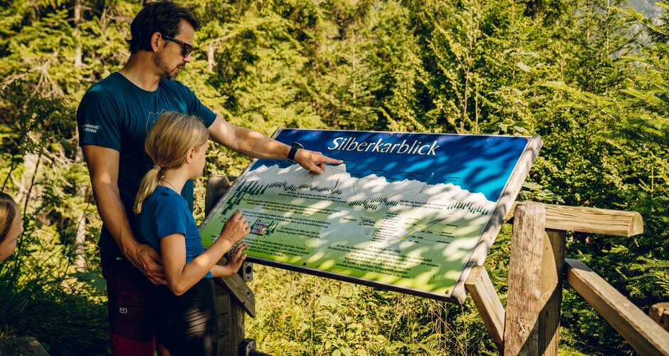 Ein Mann zeigt einem Mädchen eine Panoramatafel aus Holz am Waldrand in Ramsau am Dachstein | © TVB Schladming-Dachstein, Christine Höflehner