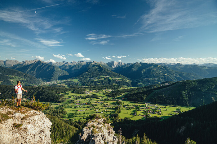 Weitblick beim Wandern in den Bergen von Schladming | © photo-austria.com, Christine Höflehner Wanderer mit rotem Rucksack steht auf Felsvorsprung und blickt über grünes Tal mit Bergen und blauem Himmel | © photo-austria.com, Christine Höflehner