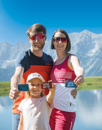 Family shows their my Ski amadé Friends Cards by the lake Smiling family holds up their my Ski amadé cards at a mountain lake with a stunning Alpine backdrop behind them.