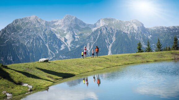 Eine Familie steht bei Sonnenschein am Ufer eines Bergsees mit klarer Spiegelung, umgeben von imposanter Alpenkulisse.