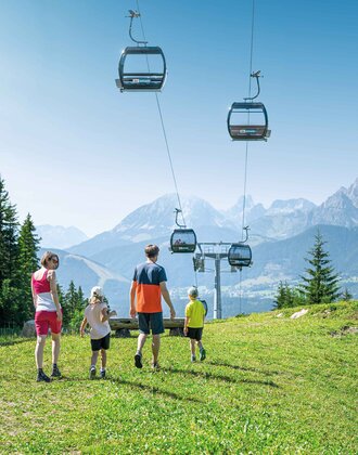 A family hikes across an alpine meadow in the sun, with a view of gondolas in the air and the impressive Dachstein mountains beyond.