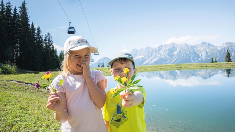 Zwei Kinder mit gepflückten Blumen stehen am Bergsee in Ski amadé, im Hintergrund die markanten Dachstein-Berge.