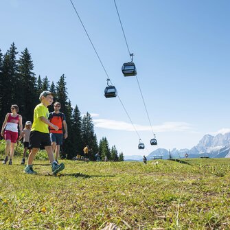 Eine Familie wandert bei Sonnenschein auf einer Almwiese unter schwebenden Gondeln mit Blick auf die Berge von Ski amadé.