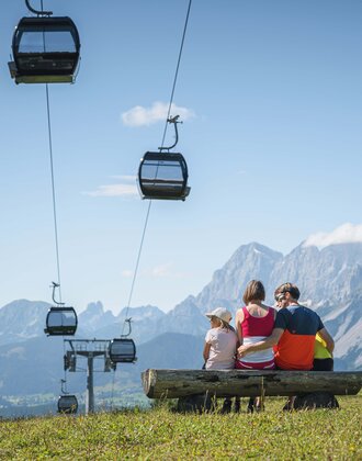 Family enjoys mountain panorama on bench beneath gondolas in Ski amadé A family sits on a wooden bench under gondolas, admiring the impressive mountain panorama in the Austrian region of Ski amadé.