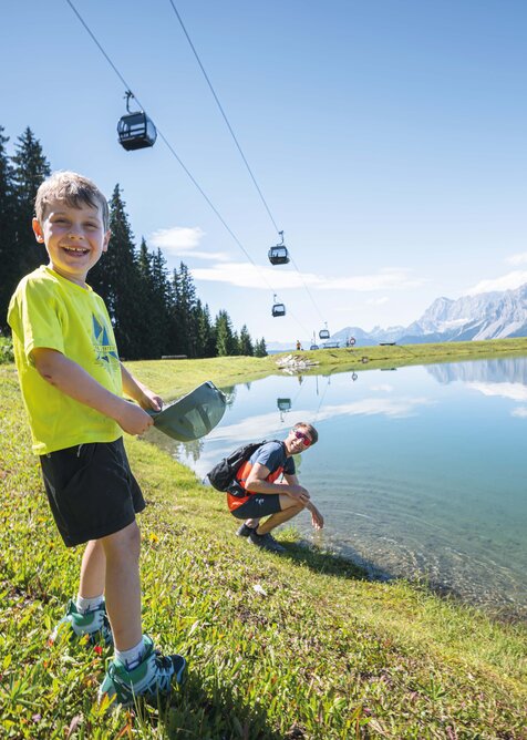 Ein Junge spielt lachend am Ufer eines klaren Bergsees, im Hintergrund kniet sein Vater – darüber fahren Gondeln vor Bergkulisse.