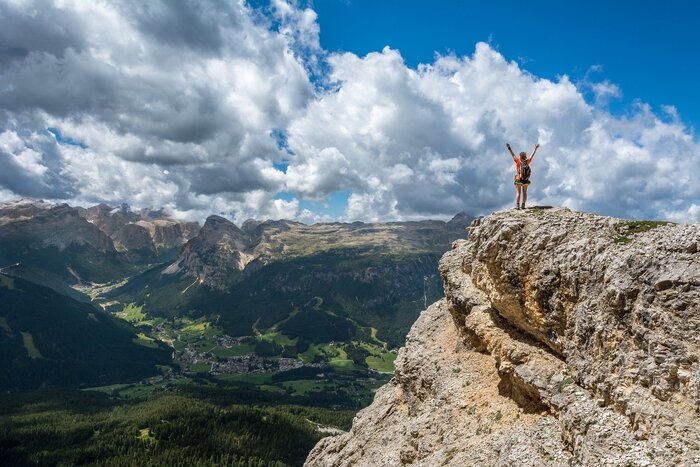 Wanderin mit erhobenen Armen auf Felsplateau mit Talblick in den Alpen Eine Wanderin mit Rucksack steht mit ausgestreckten Armen auf einem Felsplateau hoch über einem grünen Tal in den Alpen.