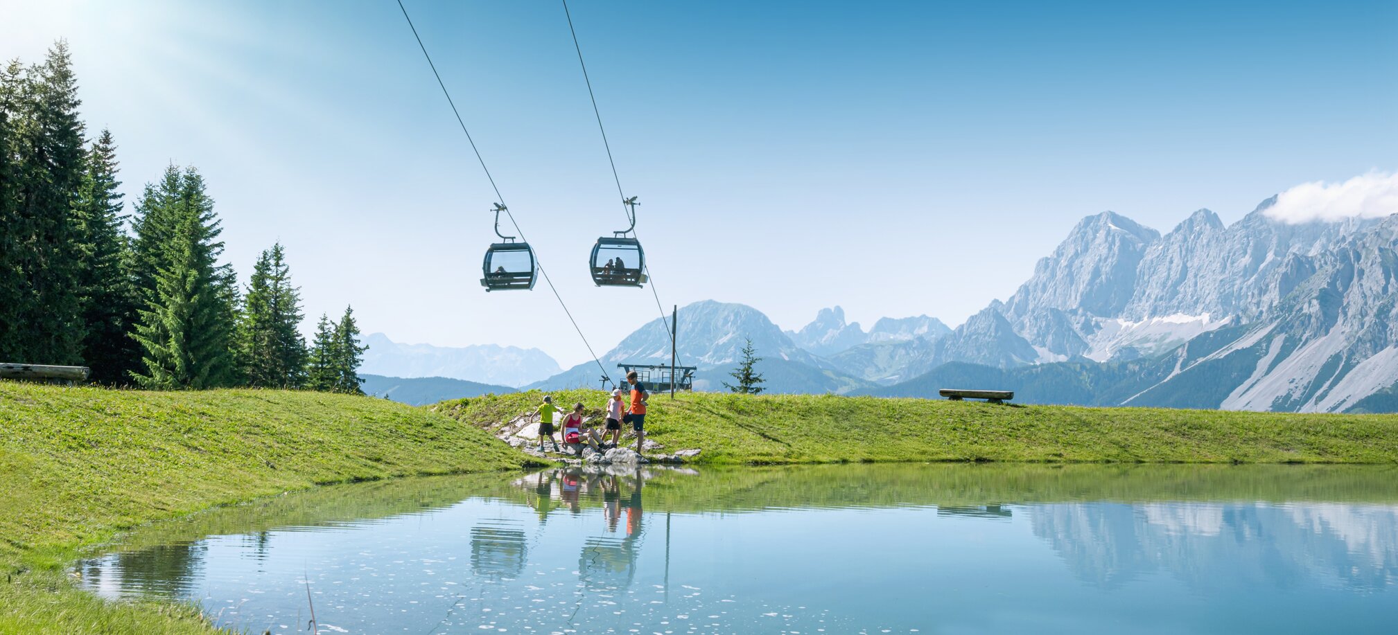 Familie am Ufer eines kleinen Bergsees unter einer Kabinenbahn, mit grünen Wiesen und Alpen im Hintergrund