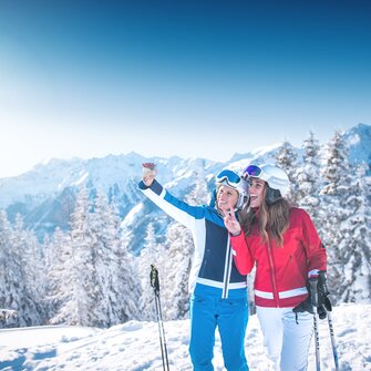 Zwei Skifahrerinnen machen ein Selfie am Pistenrand vor verschneitem Alpenpanorama, lachen und genießen die Sonne.