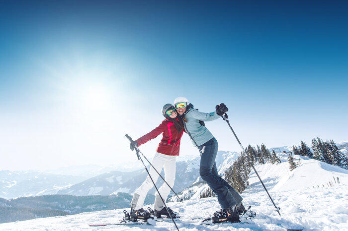 Ladies Week – Skifahren mit Freundinnen Zwei Skifahrerinnen umarmen sich auf der Piste, tragen Skibrillen und Helme, genießen das Alpenpanorama im Hintergrund.