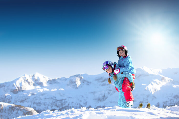 Kids playing happily in the snow Two girls in blue and red ski outfits, helmets on, play in the snow with mountains and bright sunshine behind them.