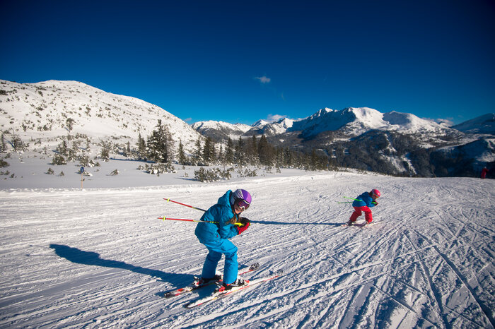 Ski amadé Minis Week – Kinder auf Skiern unterwegs Zwei Kinder in bunter Skikleidung fahren auf Skiern die Piste hinab, genießen die verschneite Alpenlandschaft.