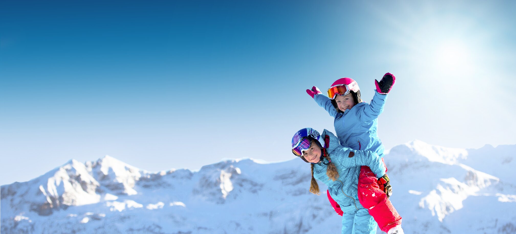 Two girls in blue ski outfits play and laugh in the snow with snowy mountains in the background, one raising hands.