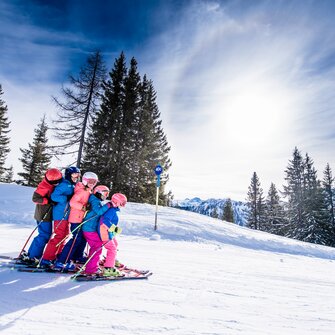 Sechs Kinder in bunter Skikleidung fahren eng zusammen auf Skiern die Piste hinab, lachen und genießen den Schnee.