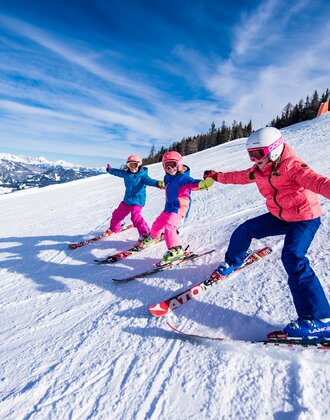 Drei Kinder fahren mit bunten Skianzügen nebeneinander auf der Piste, halten sich an den Händen und lachen im Schnee.