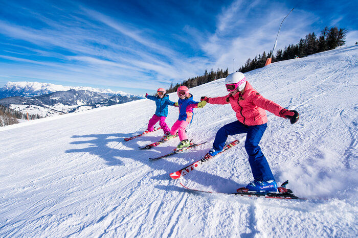 Ski amadé Minis Week – kids' ski fun on the slopes Three kids in colorful ski outfits ski side by side on the slope, holding hands and laughing in the snow.