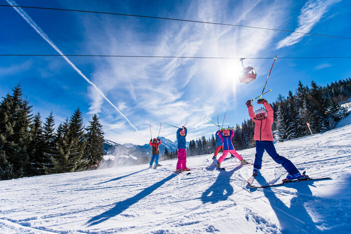 Eine Gruppe von Kinder in bunten Skianzügen heben Skistöcke auf der Piste in die Luft, lächeln und genießen die Sonne.