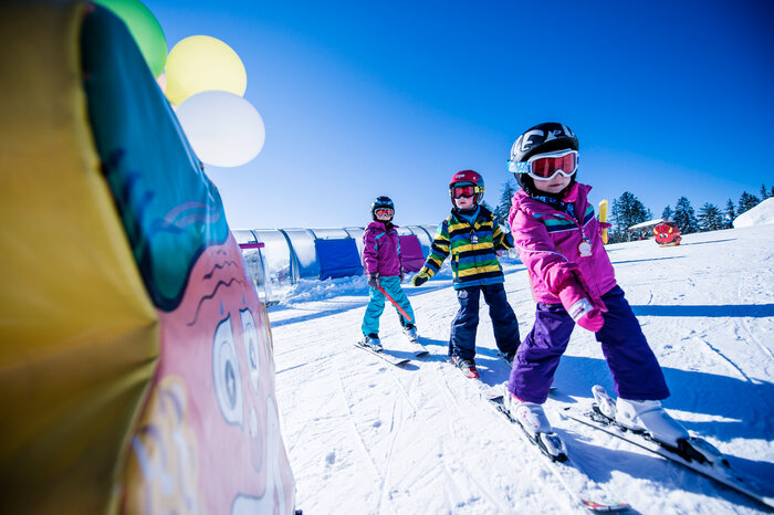 Kinder lernen Skifahren mit Spaß Drei Kinder in bunten Skianzügen fahren auf einer sonnigen Skipiste zwischen Skischule-Aufbauten, lachend und motiviert.