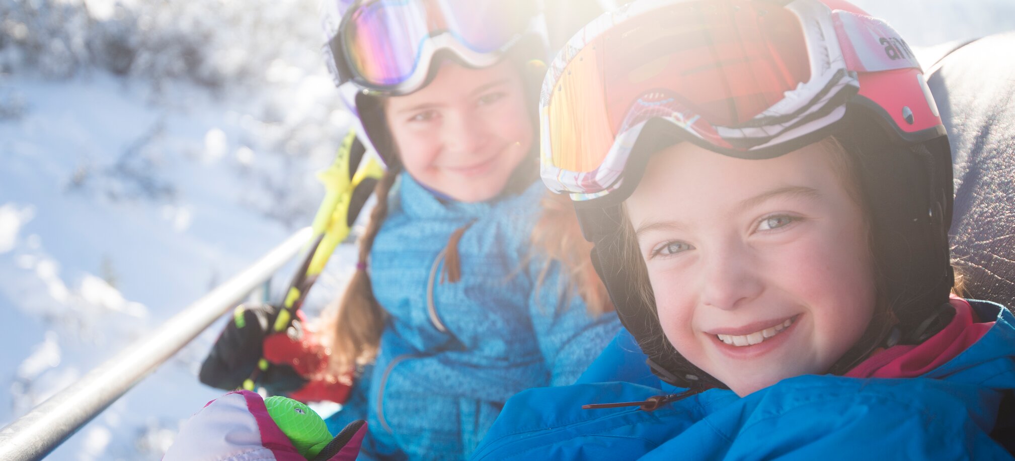 Zwei Kinder in bunten Skianzügen und Helmen sitzen lachend im Skilift, genießen die Sonne und Winterlandschaft.