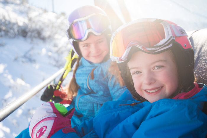 Ski amadé Minis Week – lachende Kinder im Skilift Zwei Kinder in bunten Skianzügen und Helmen sitzen lachend im Skilift, genießen die Sonne und Winterlandschaft.