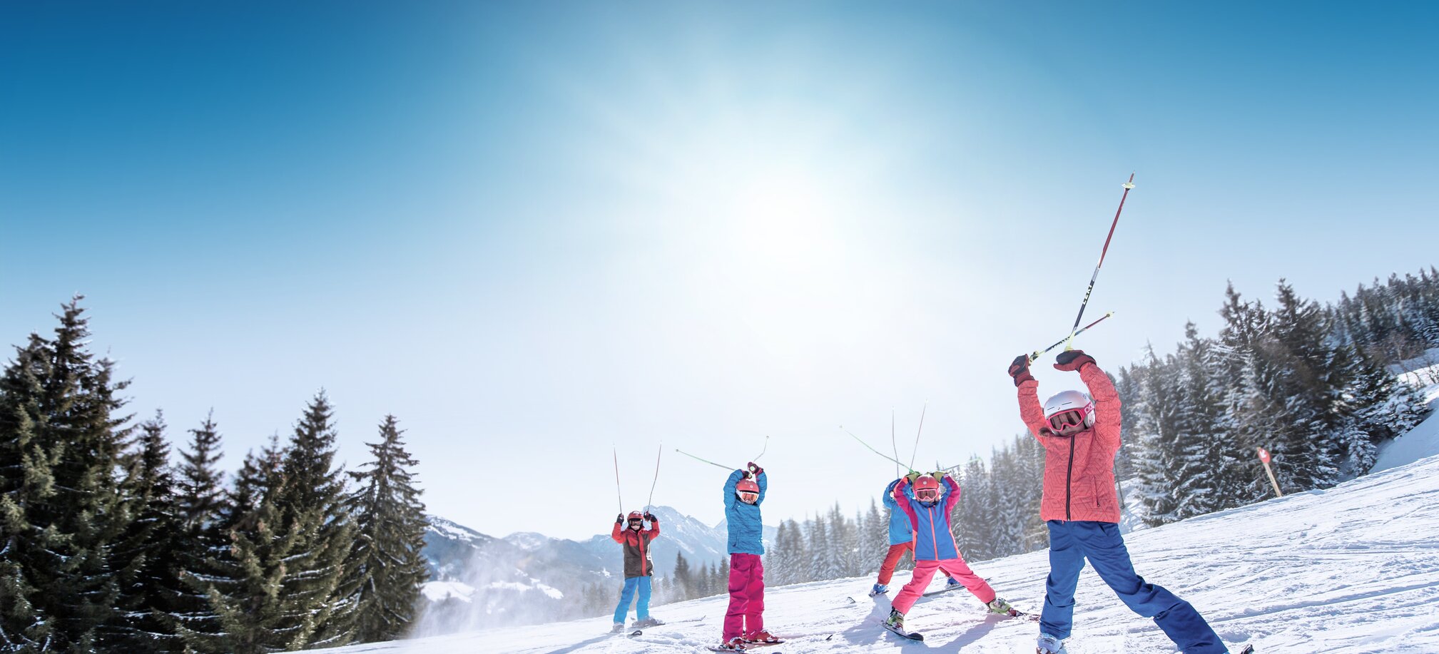 Kinder in bunter Skikleidung heben Skistöcke freudig in die Luft, vor verschneiten Bergen und Pisten.