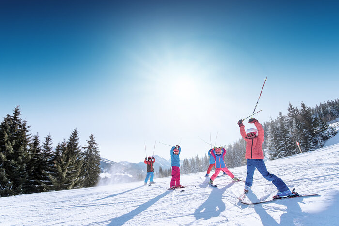 Ski amadé Minis Week – kids cheer in front of Alps Kids in colorful ski outfits raise ski poles happily in the air, with snowy mountains and slopes in the background.
