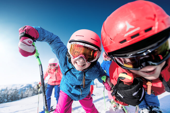 Ski amadé Minis Week – fun in the snow Kids with colorful helmets and ski goggles skiing downhill, smiling under the sun, snowy mountains in the background.