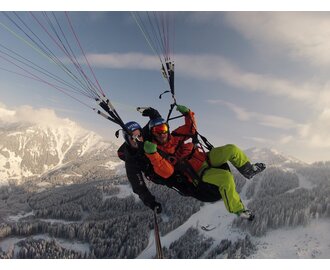 Zwei Menschen gleiten beim Tandem-Paragliding über eine schneebedeckte Berglandschaft, umgeben von Winterpanorama.