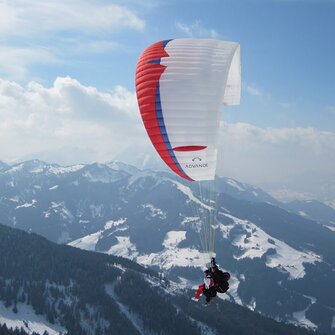 Paragleiter mit rot-blau-weißem  Schirm fliegt über von Wald und schneereichen Pisten bedeckte Berge
