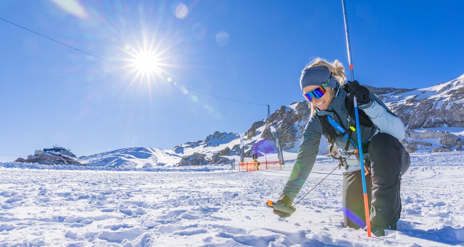 Frau kniet im Schnee mit Sonde und LVS-Gerät, Sonnenschein und Berge im Hintergrund.  | © René Eduard Perhab