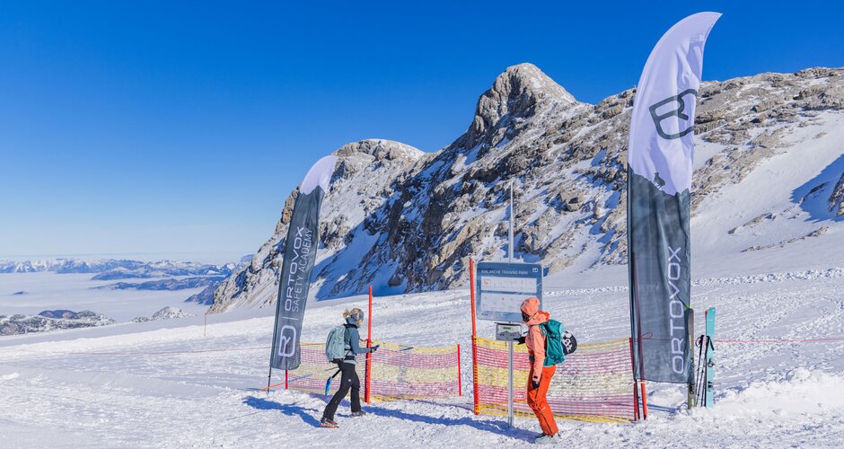Zwei Wintersportler vor einem Trainingsbereich im Schnee mit Fahnen, Zaun und Gebirgskulisse am Dachstein.  | © René Eduard Perhab