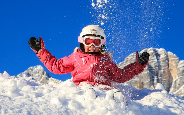 Schneeabenteuer für Kinder in Ramsau am Dachstein Kind in pinker Skibekleidung spielt lachend im Schnee vor blauer Himmel und Bergen.