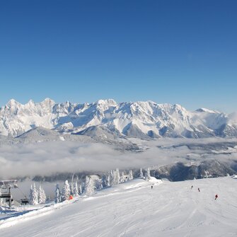 Blick auf verschneite Piste, Skifahrer und beeindruckendes Bergpanorama im Hintergrund.