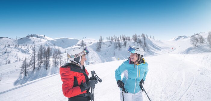 Two female skiers stand on the slope and laugh together under a bright and sunny sky