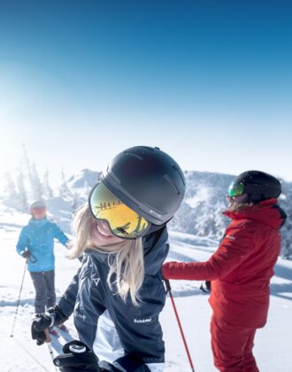 Frau mit Helm und Skibrille lacht in die Kamera, im Hintergrund zwei Skifahrer im Schnee