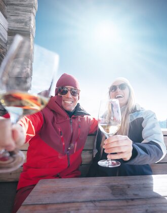 Zwei Skifahrer stoßen mit Weißwein auf Holzbank vor Hütte in Ski amadé an, Sonne und Berge im Hintergrund
