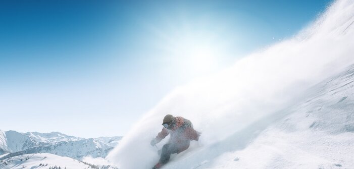 Snowboarder fährt im Tiefschnee eine steile Hangflanke hinab, Pulverschnee spritzt auf