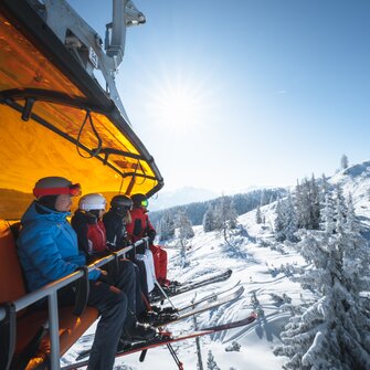 Group of skiers with helmets and goggles riding a chairlift with an orange cover over a snowy winter landscape. | © Ski amadé