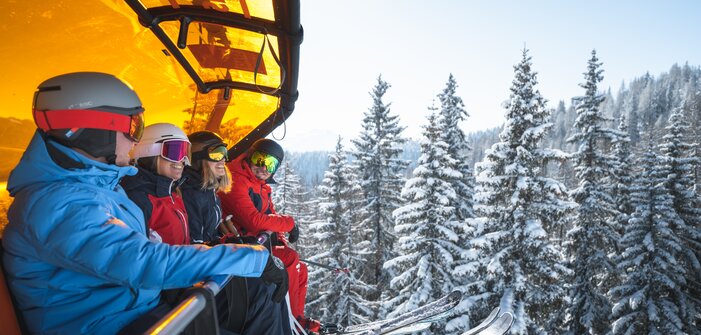 Vier Skifahrer mit Helmen und Skibrillen sitzen auf einem Sessellift mit orangener Haube über verschneiten Tannen. | © Ski amadé