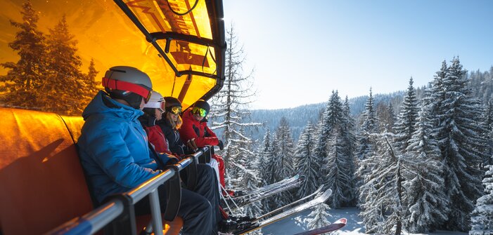 Vier Skifahrer mit Helmen und Skibrillen sitzen auf einem Sessellift mit orangener Haube über verschneiten Bäumen. | © Ski amadé