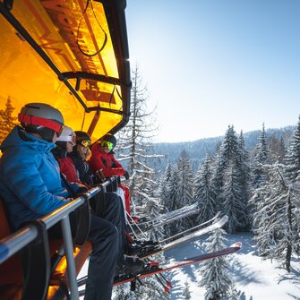 Four skiers wearing helmets and goggles sit on a chairlift with an orange cover above snow-covered trees. | © Ski amadé