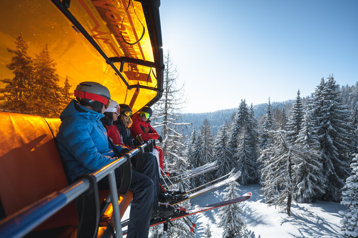 Skiers on Chairlift with Orange Weather Cover | © Ski amadé Four skiers wearing helmets and goggles sit on a chairlift with an orange cover above snow-covered trees. | © Ski amadé